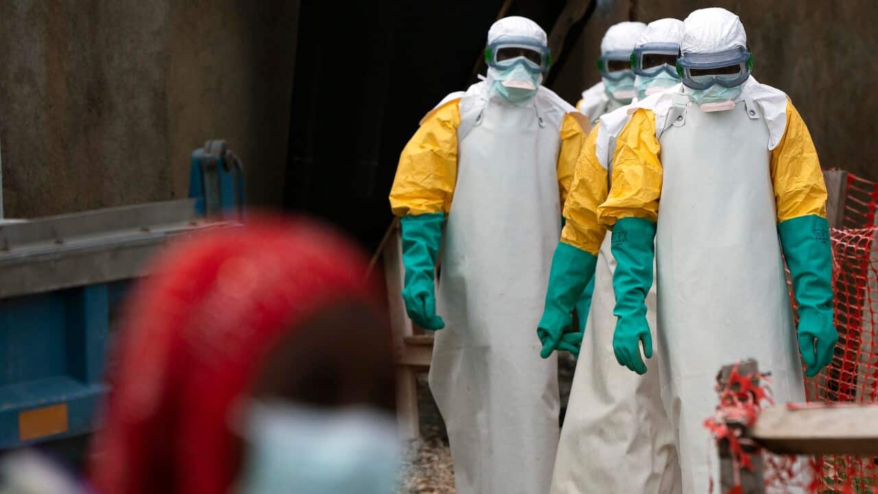 Four people in protective hazmat suits, gloves and goggles walk towards a person in a mask.