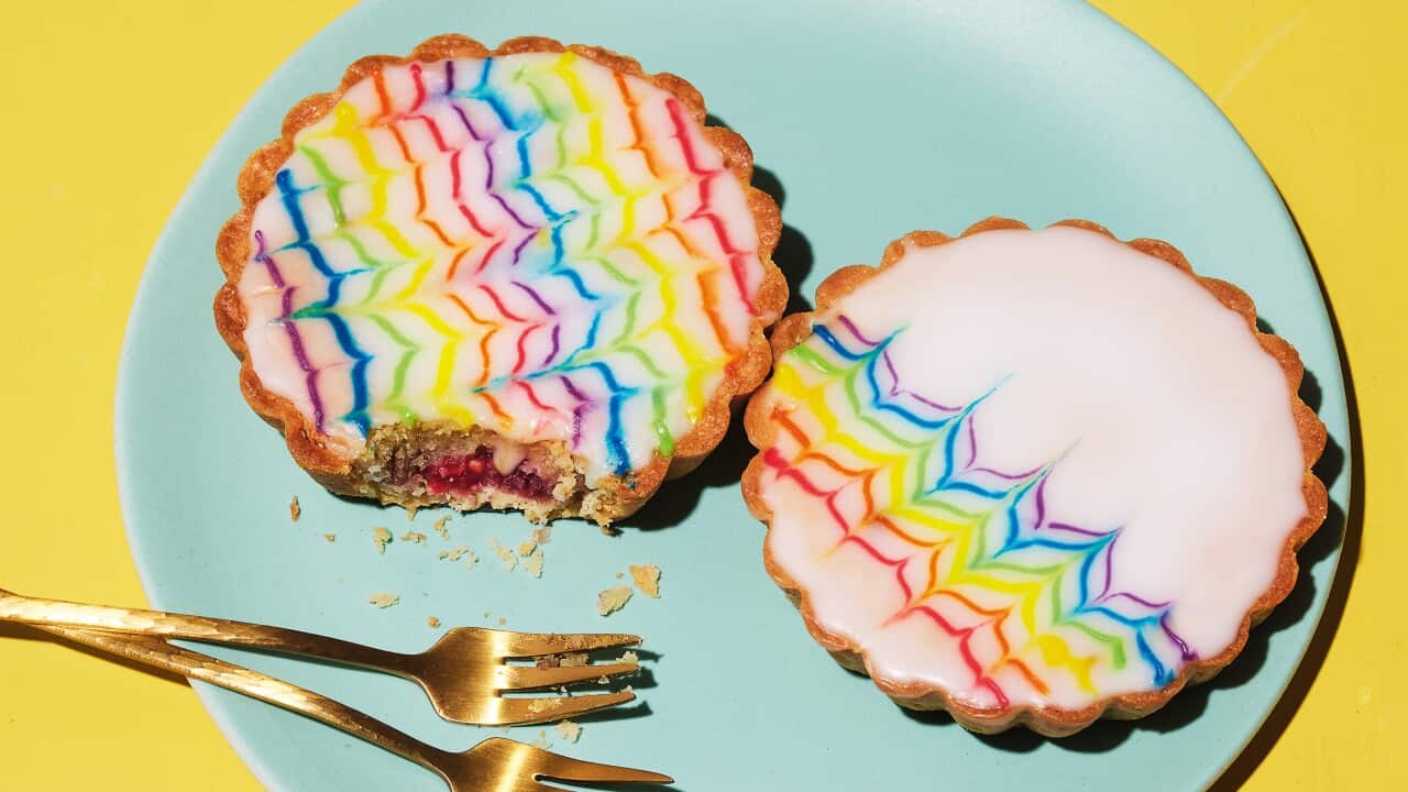Two small round tartletes with fluted pastry edges sit on a blue plate on a bright yellow background. Each tart is decorated with white icing with a feathered pattern in rainbow colours.