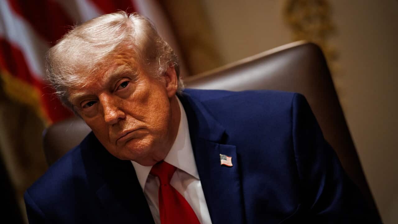 US President Donald Trump sits in a high-backed chair wearing a dark blue suit, white shirt and red tie, with a US flag pin on his lapel. He is leaning slightly to one side, with a blurred American flag and an indoor setting visible in the background.
