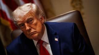 US President Donald Trump sits in a high-backed chair wearing a dark blue suit, white shirt and red tie, with a US flag pin on his lapel. He is leaning slightly to one side, with a blurred American flag and an indoor setting visible in the background.