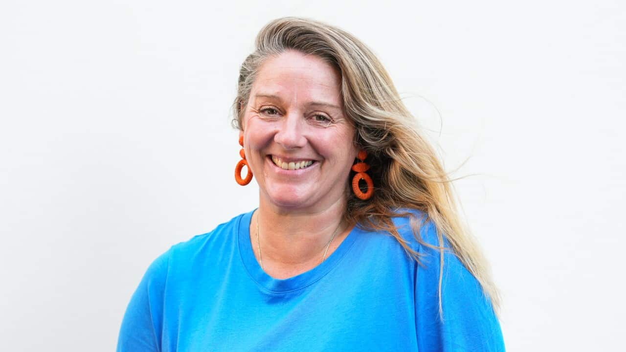 a woman with long blonde hair, red earrings, a blue tshirt and red and blue pants poses in front of a white background