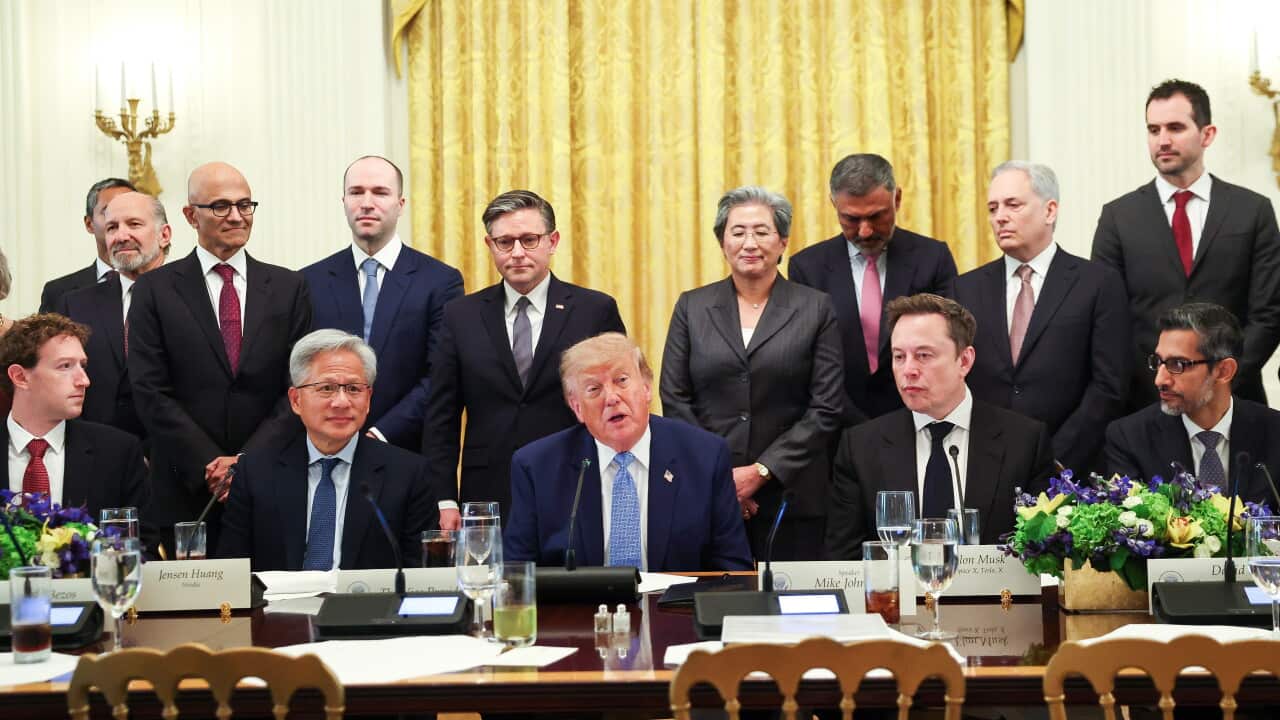 Group of business and government leaders seated and standing around a conference table during a formal meeting, with microphones, nameplates and floral arrangements visible.
