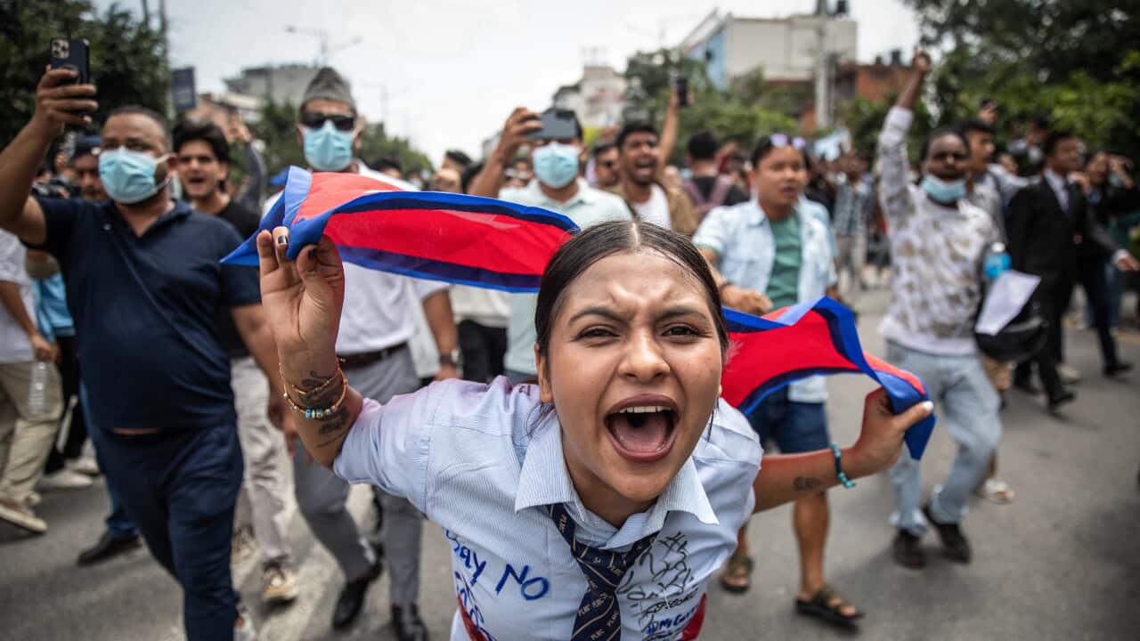 A young woman shouting and holding the Nepali flag leads a crowd of protesters through a street, with "Say No" and other slogans written on her shirt.