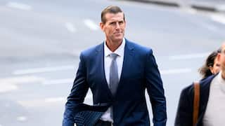 A tall middle-aged man in a blue suit and tie walks on a city street. His jacket appears to be caught by the wind.