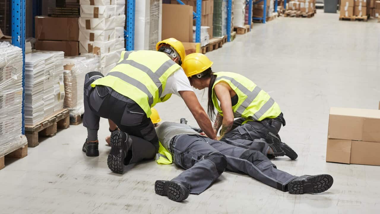 A person collapsed on the floor of a warehouse with two people kneeling over them