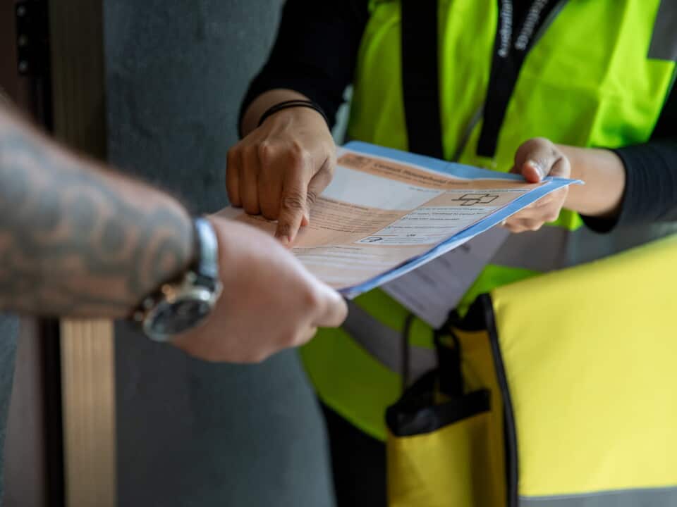 A woman wearing a fluorescent yellow vest hands a man an orange Census form. He wears a watch and has tattoos on his forearm.