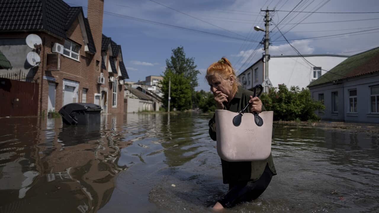 Elderly woman holds one hand to her face, her brow furrowed, as she walks through floodwaters while carrying a handbag.