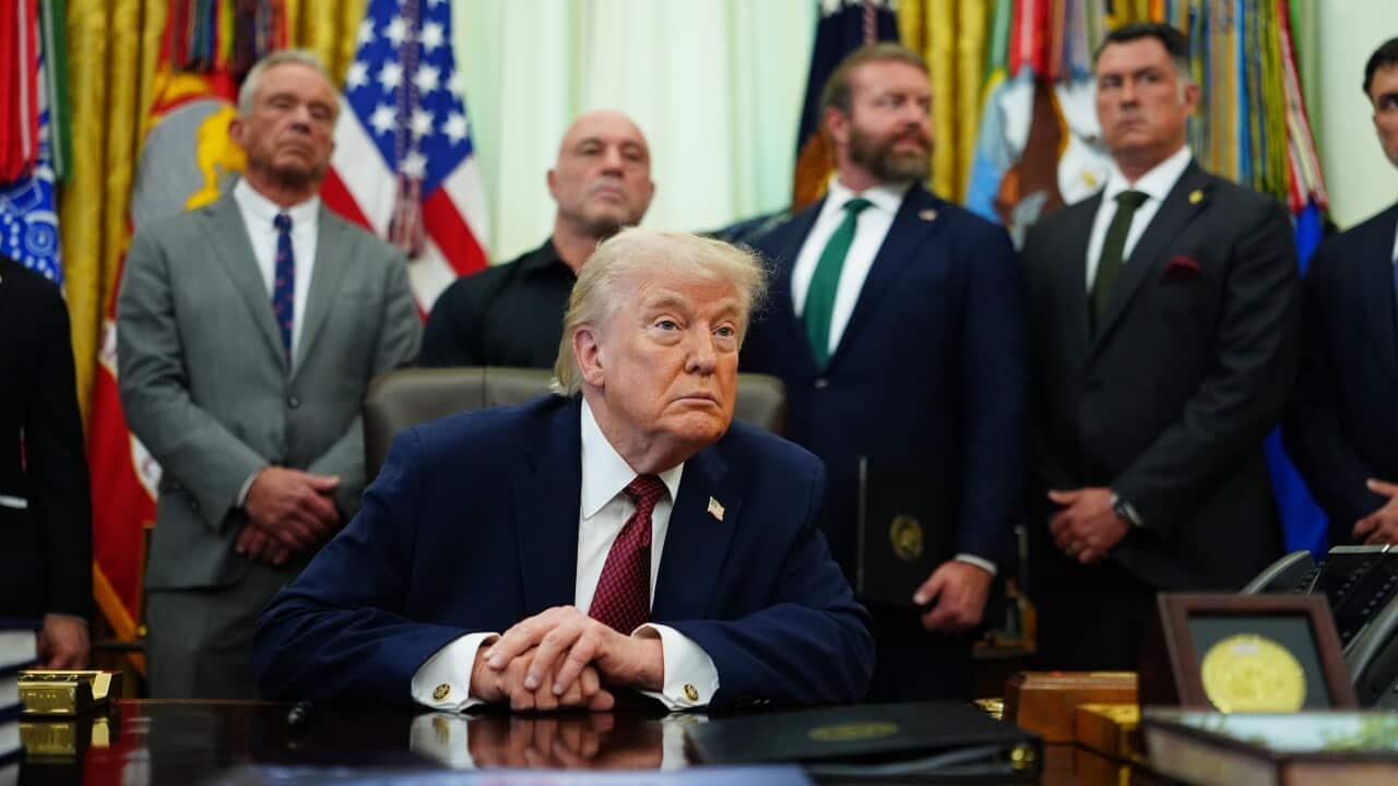US President Donald Trump in the Oval Office, sitting behind his desk, flanked by men in suits