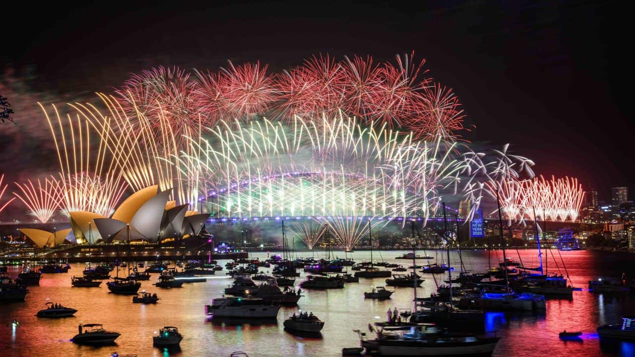 Fireworks illuminate the skyline, with a bridge and nearby buildings, as boats sail in the foreground.