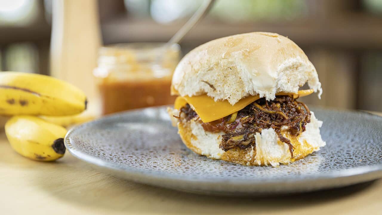 A soft white bun, filled with a pulled brown filling and a slice of cheese, sits on a speckled grey plate. Two bananas and a jar of sauce can be see in the background.