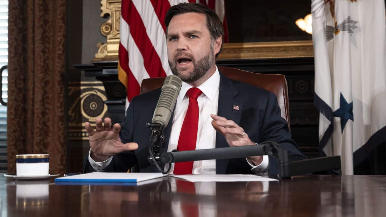 JD Vance, a Caucasian man with a beard and a suit, sits at a desk, speaking into a microphone. He gestures with his hands, with an American flag visible in the background.
