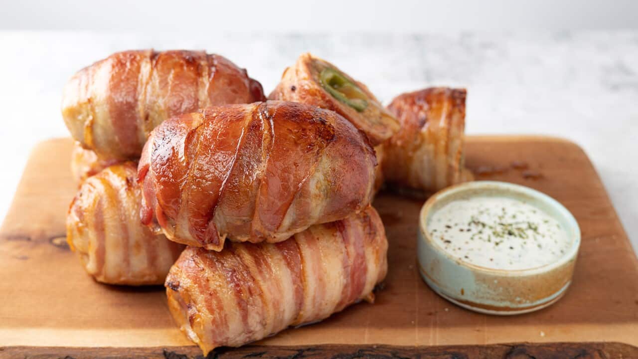 Five bundles, wrapped in strips of bacon, and a small pot with white sauce, sit on a chunky wooden board.