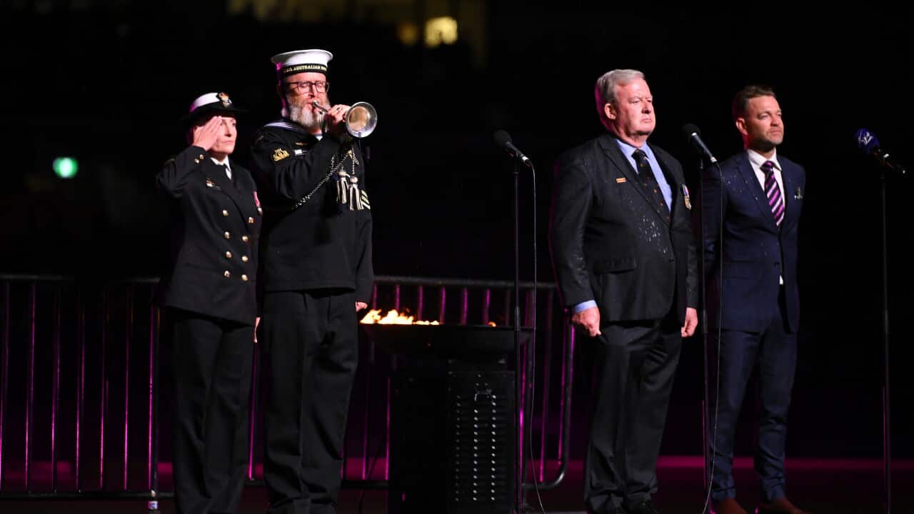 Four people stand on a stage. Two are wearing formal military attire and one blows a trumpet.