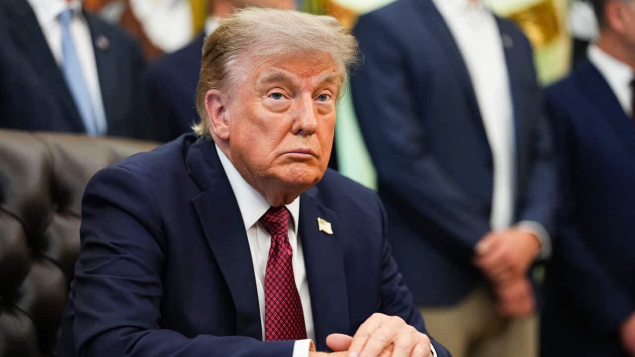US President Donald Trump sits at a desk in the White House with his hands clasped as officials stand behind him during a meeting.