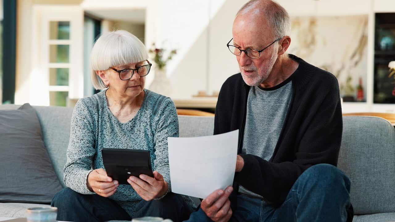 A senior couple sitting on a couch in front of a table with papers on it. The man is holding up a piece of paper that they are both looking at