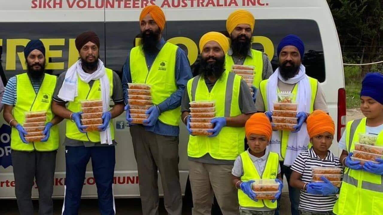 A group of men and boys wearing turbans and high visibility vests stand in front of a van, holding containers of food