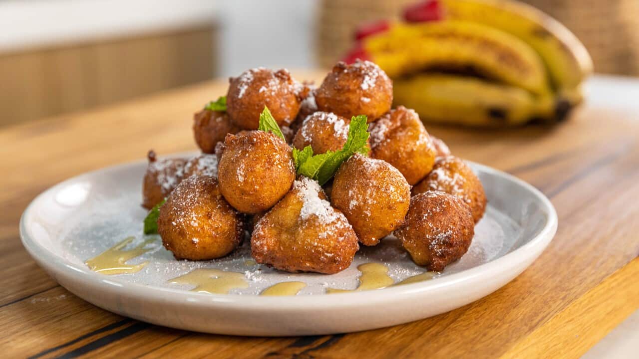 A pile of golden roundish fritters sit on a wide white plate. The fritters have been dusted with icing sugar and the plate drizzled with syrup. Bananas can be seen in the background.