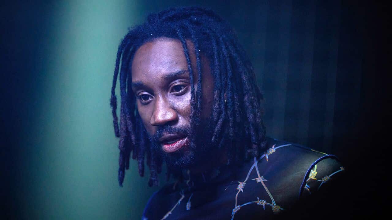 A man with dreadlocks in a top with a barbed wire pattern stares ahead, slightly downward.