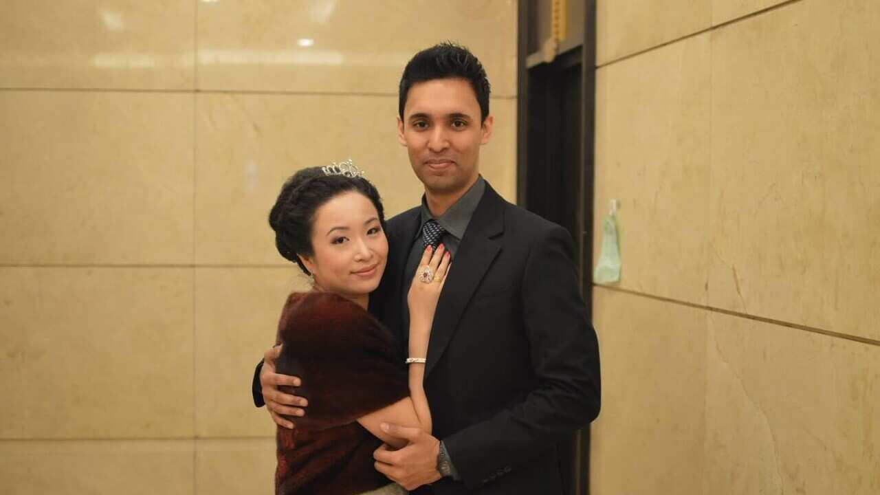 A woman wearing a tiara hairpiece and a dress with a vest coat places her hand on the chest of a man wearing a black suit.