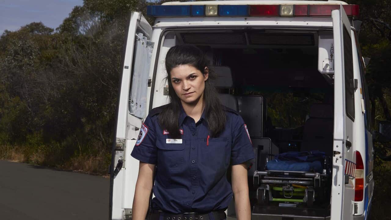 A young woman with long dark hair, in a paramedic's uniform and blue disposable gloves, stands in front of the open rear doors of an ambulance.