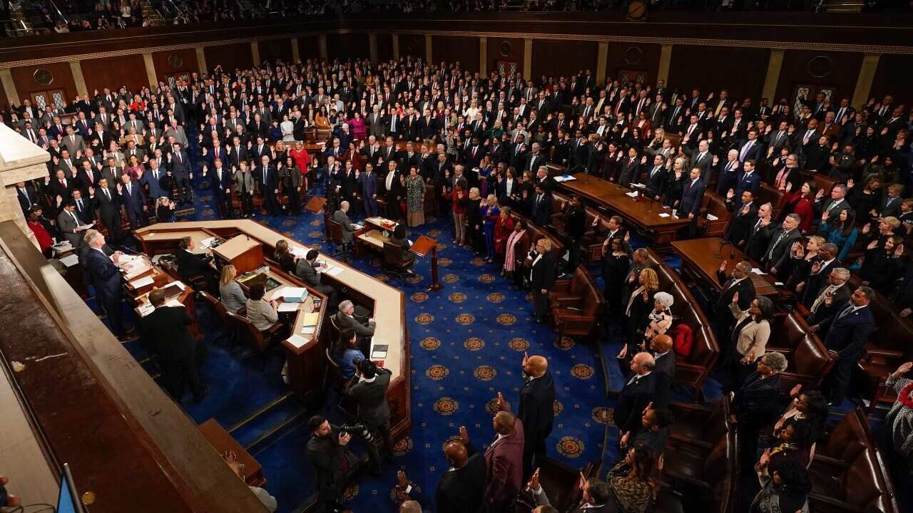 A room of people wearing suits stand as they are sworn in as members of the US House of Representatives.