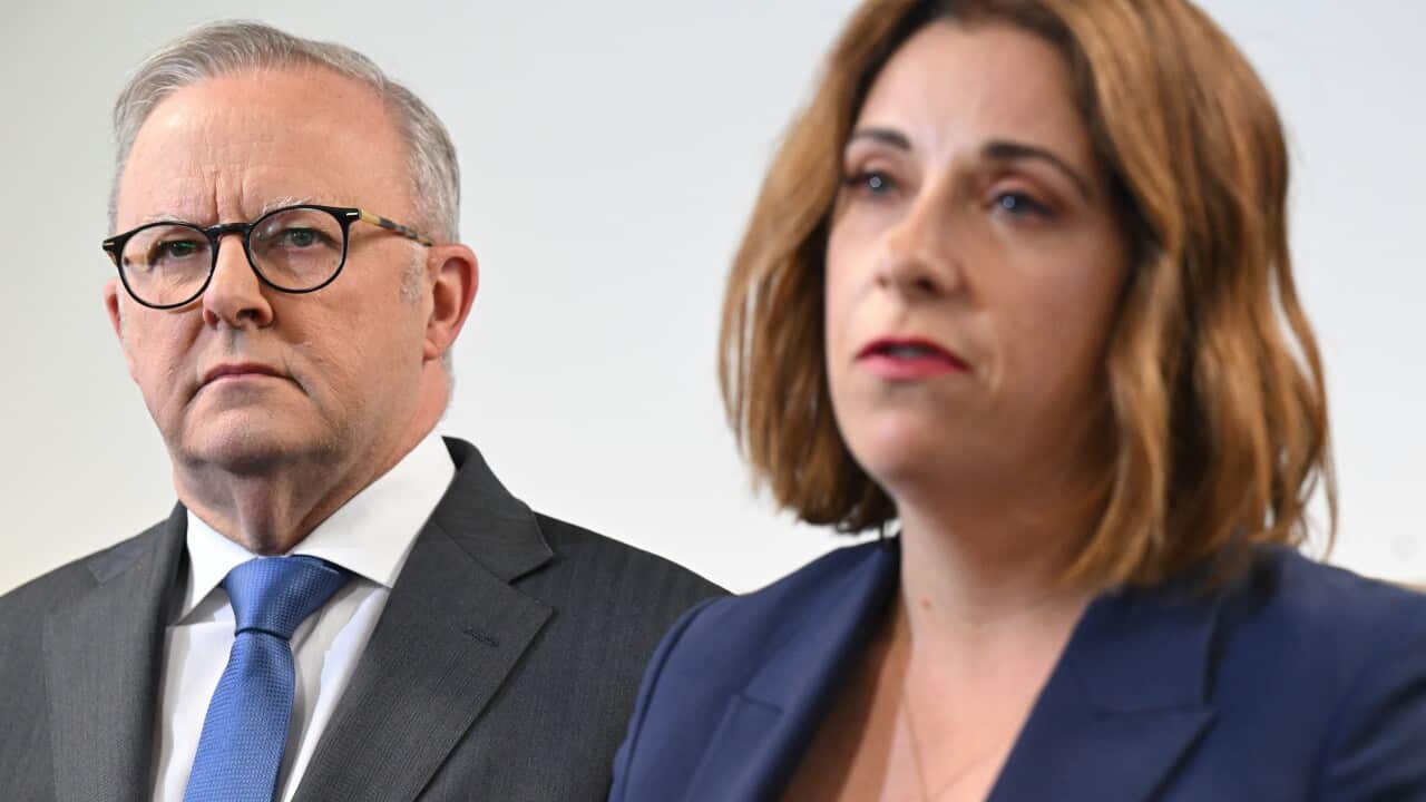 A man in focus in a dark suit and glasses standing behind a woman with short auburn hair in the foreground