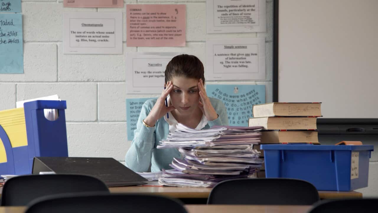 Schoolteacher at desk staring at piled exercise books, hands to head