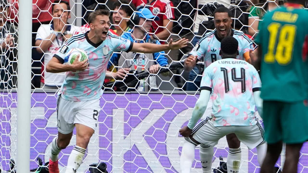 Three footballers dressed in light blue celebrate a goal in front of an excited crowd.