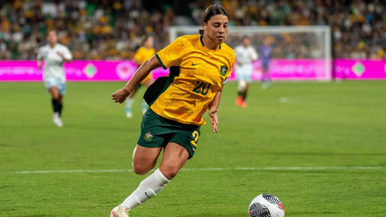 A woman in a yellow football shirt and green football shorts running next to a ball.