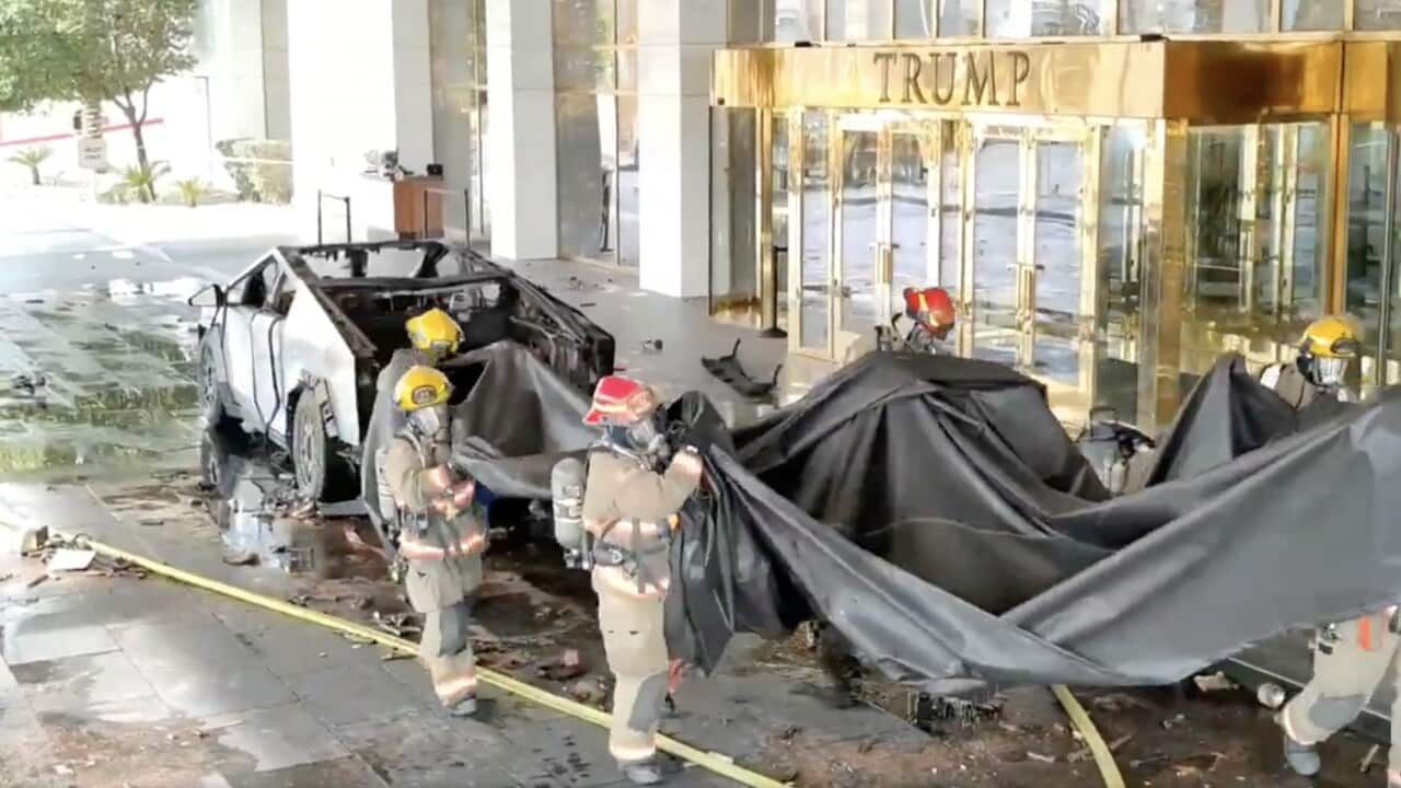 Several firefighters remove a black tarp from the charred husk of a Tesla Cybertruck sitting in front of a hotel entrance with "Trump" emblazoned above the entrance