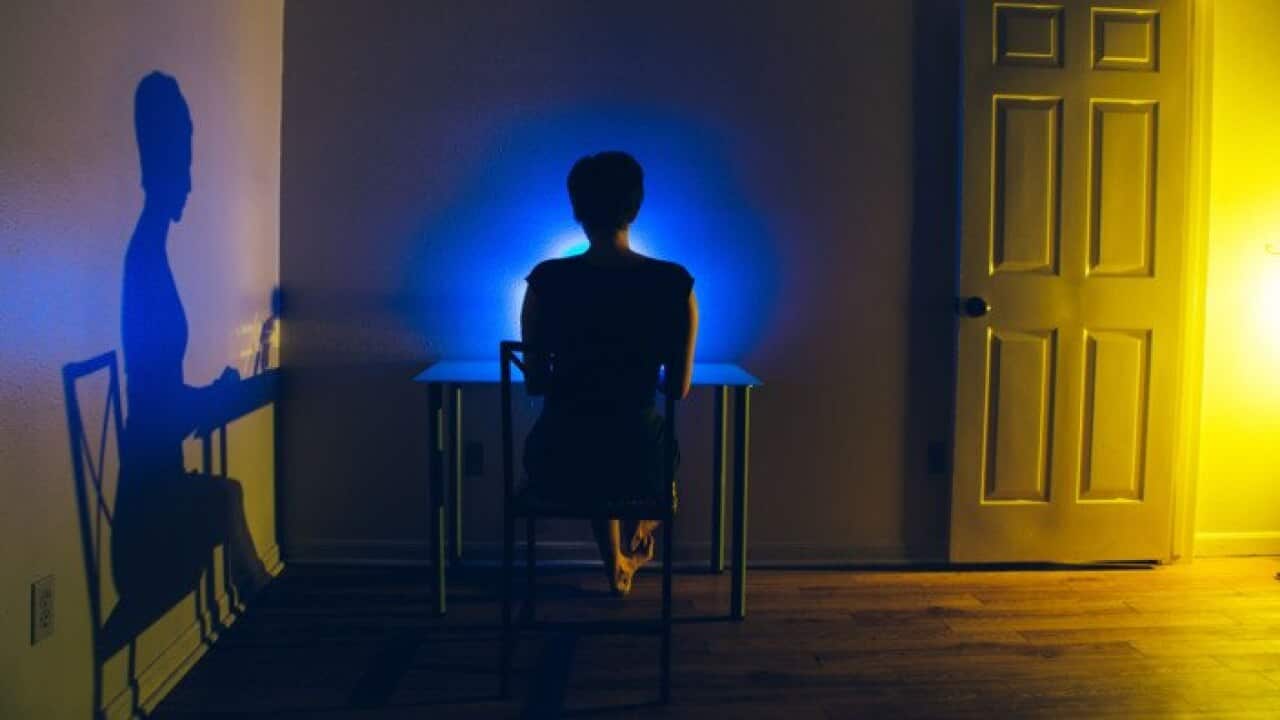 woman sitting at desk in a dark room. Open door allows amber light in which produces a shadow of the woman on the wall. Blue light from computer illuminates woman's head.