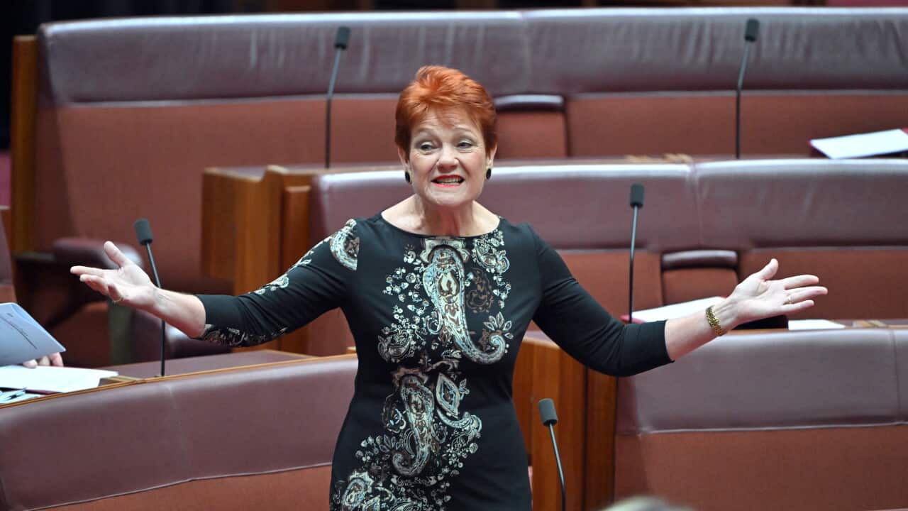 A red-haired woman gestures with outstretched arms while speaking in a parliamentary chamber.