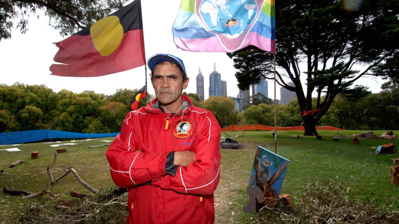 A man wearing a red jacket. Behind him is the Aboriginal flag.