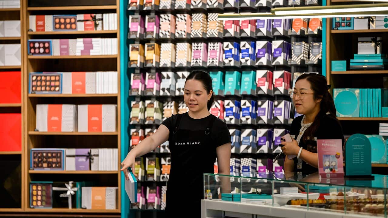 A young retail worker directs a customer to some nearby products. They are in a dessert and chocolate shop.