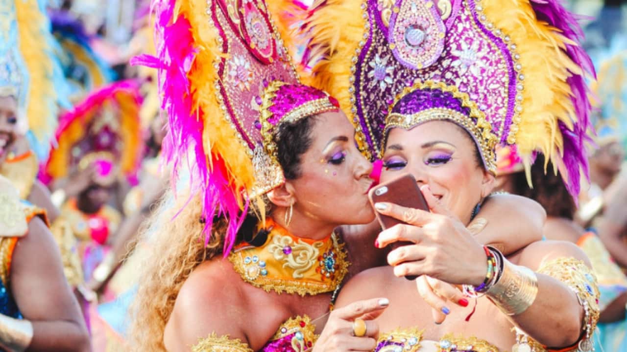 Two women Carnival dancers pose for a selfie as one kisses other on the cheek.