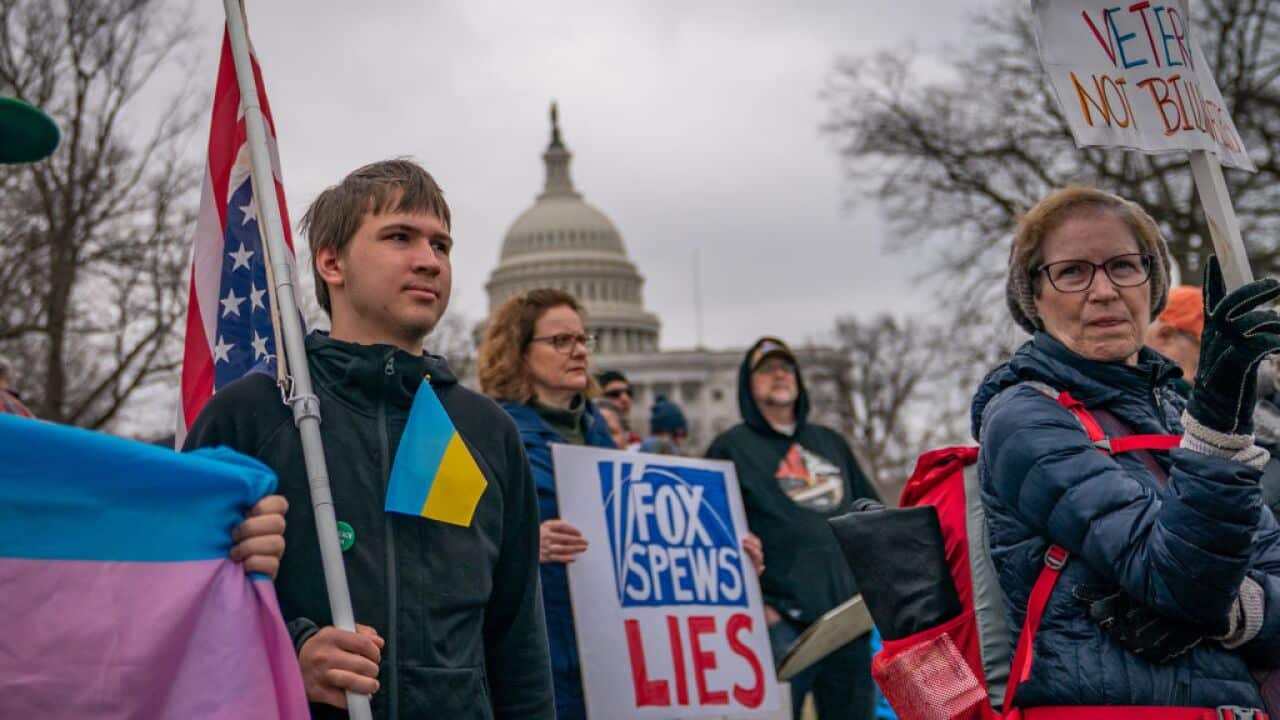 A crowd of people holding up signs protesting Fox News, in front of the US Capitol Building. A man on the left has a Ukrainian flag in his breast pocket and is holding a US flag. A woman next to him has a placard that reads 'Fox spews lies' and another on the far right holds a sign reading: Veterans, not billionaires.