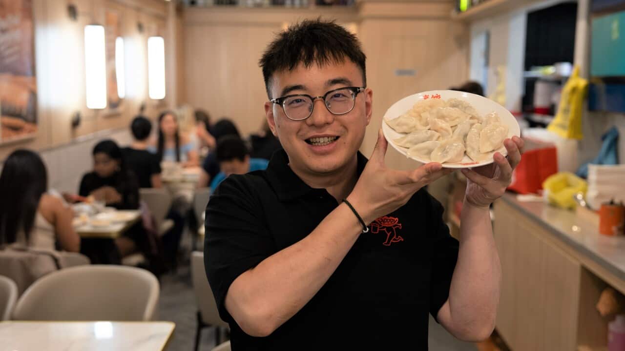 A man wearing glasses and a black t-shirt stands in a restaurant holding a plate of dumplings.