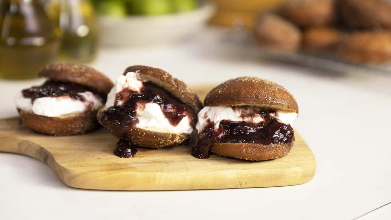 Three doughnuts, spilt and filled with cream and a jammy dark red sauce, sit on a wooden board.