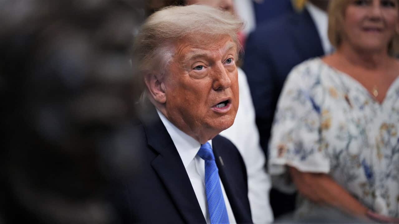 Donald Trump speaks in the Oval Office with people in formal attire gathered behind him.