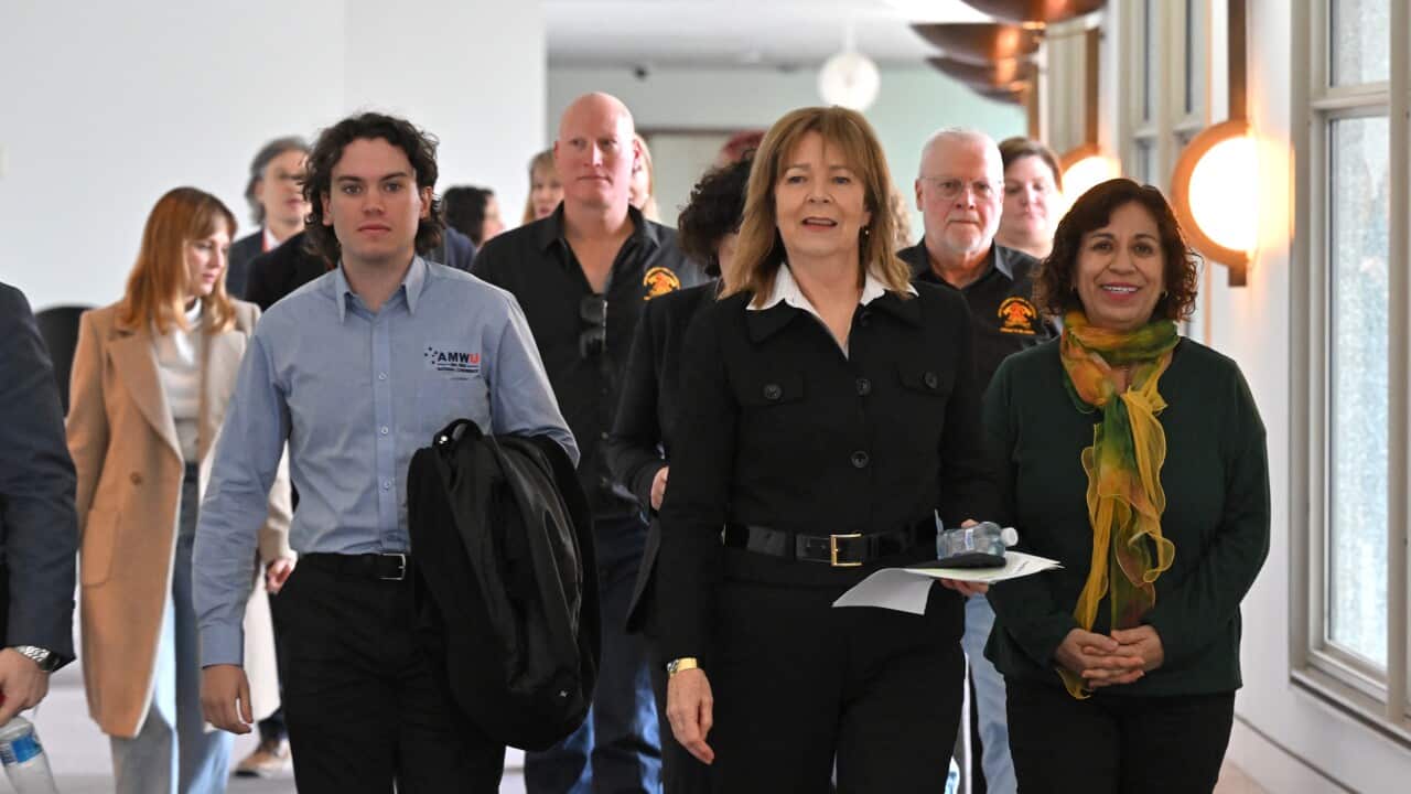 A group of people walk down a hallway, in the lead is a woman in a black shirt and pants carrying a piece of paper