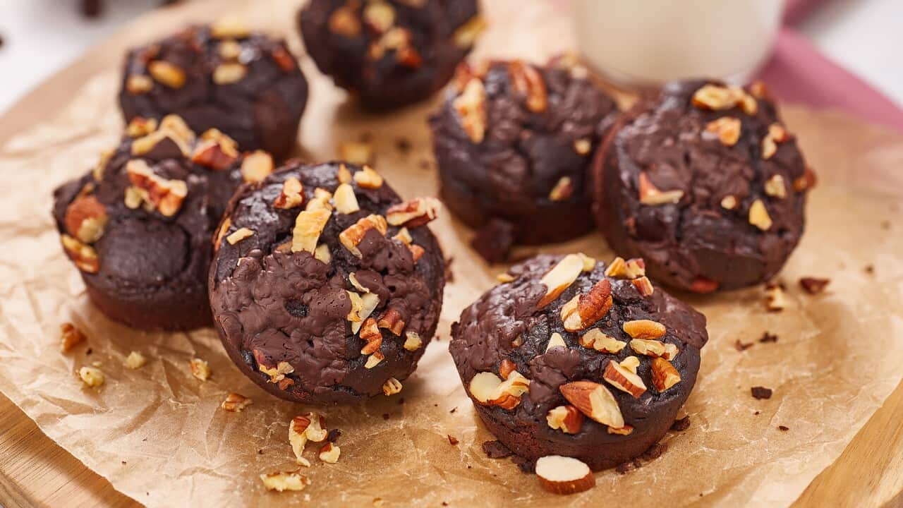 Seven muffin-shaped chocolate brownies, topped with chopped nuts, sit on a piece of baking paper on a wooden board.