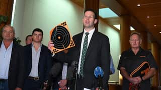 A man in a suit and green patterned tie speaks at a press conference microphone while holding up a bright orange and black target sheet, flanked by several other men indoors.