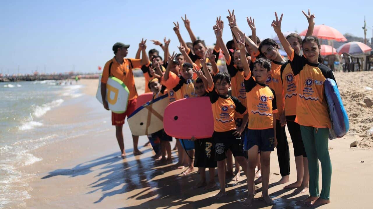 A group of children and one adult instructor stand barefoot on a sandy beach near the ocean, all raising their hands to make V for peace signs. The children are wearing matching orange and black tops and several are holding bodyboards.