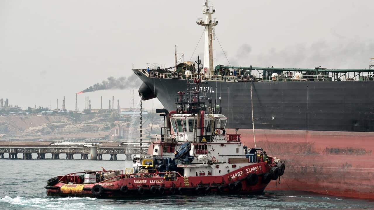 A smaller boat alongside an oil tanker at a sea port with an oil refinery in the background