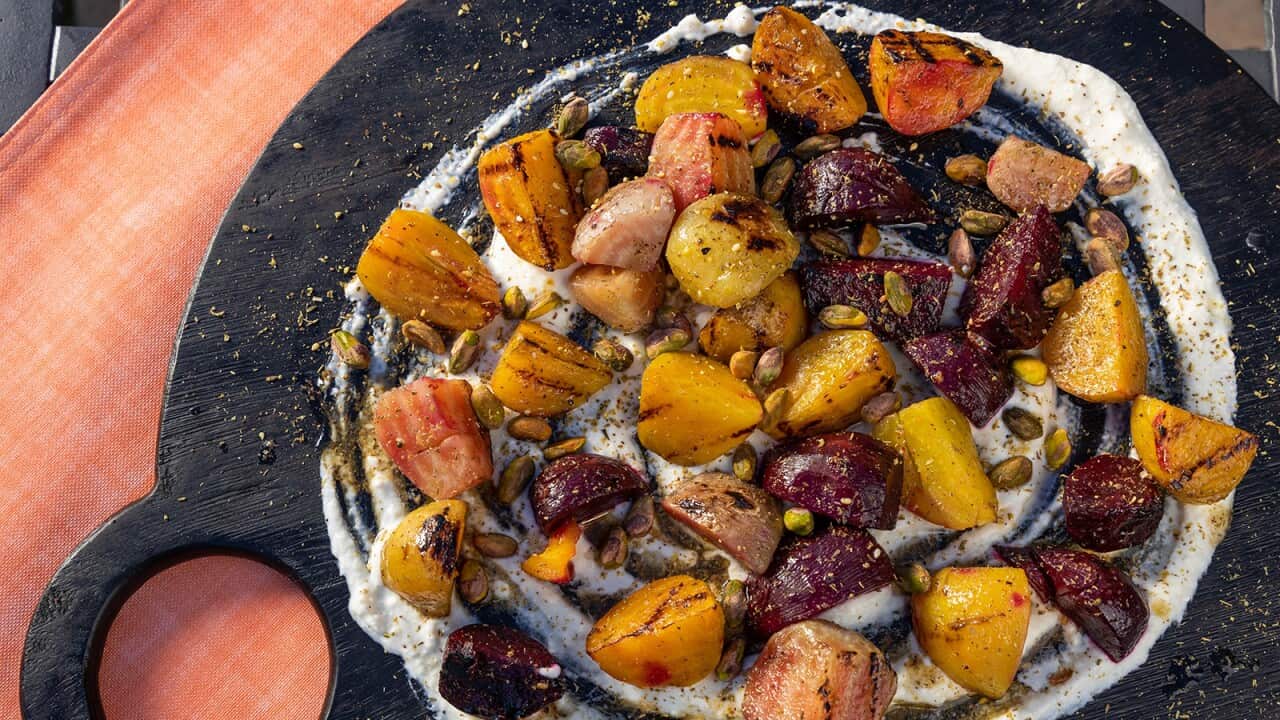 On a round, dark board with a cut-out handle coloured beets are scattered over a round creamy base. The beets are sprinkled with pistachios.