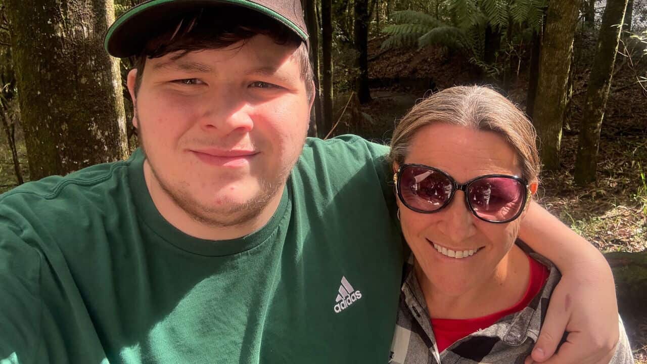 a teenage boy in a green top and cap has his arm around his mum who is wearing sunglasses. They're standing in front of a bushland area.