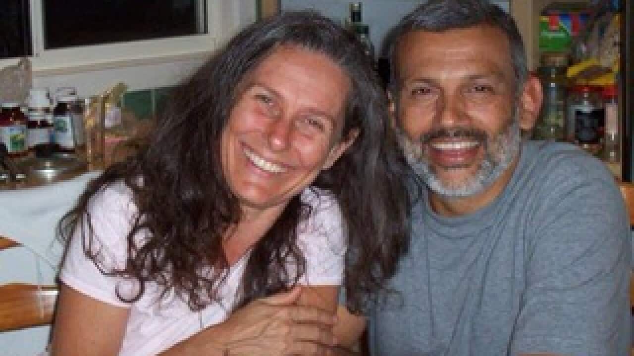A woman and man sitting in a kitchen embrace as they smile at the camera.