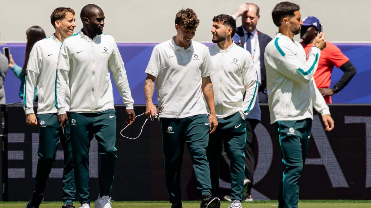 Socceroos players walk across the pitch in training gear ahead of a match, with one player holding up a phone and others talking and smiling.
