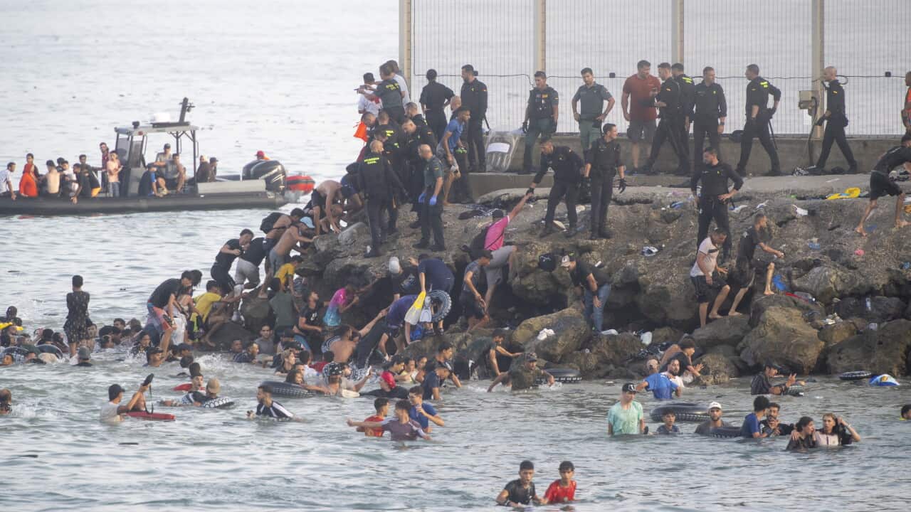 Dozens of migrants in the sea next to a rock breakwall. Uniformed police are standing on the breakwall, and a small boat carrying more migrants is in the background.