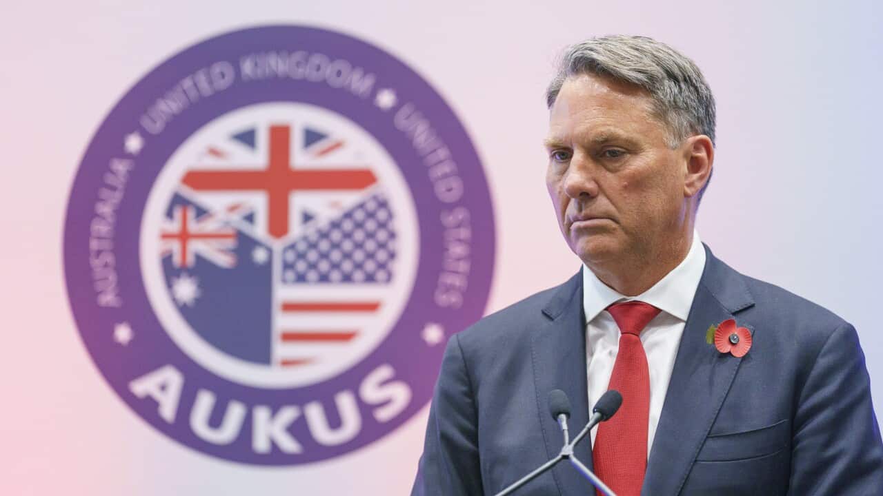Richard Marles standing behind a microphone at a podium. Behind him is an AUKUS logo with the Australian, American and British flags.